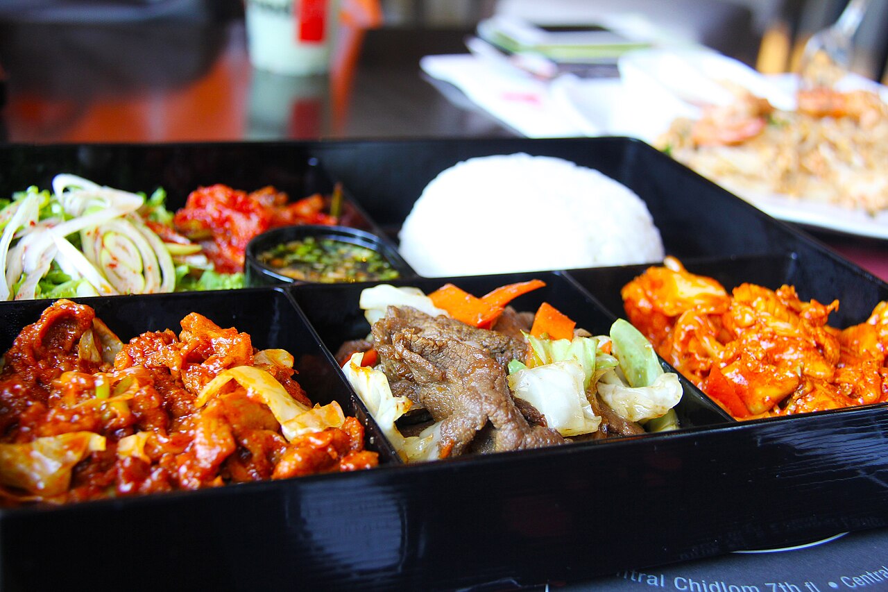 검은 칸막이 도시락 용기에 흰밥과 고기, 채소, 붉은 양념 반찬이 나뉘어 담긴 모습